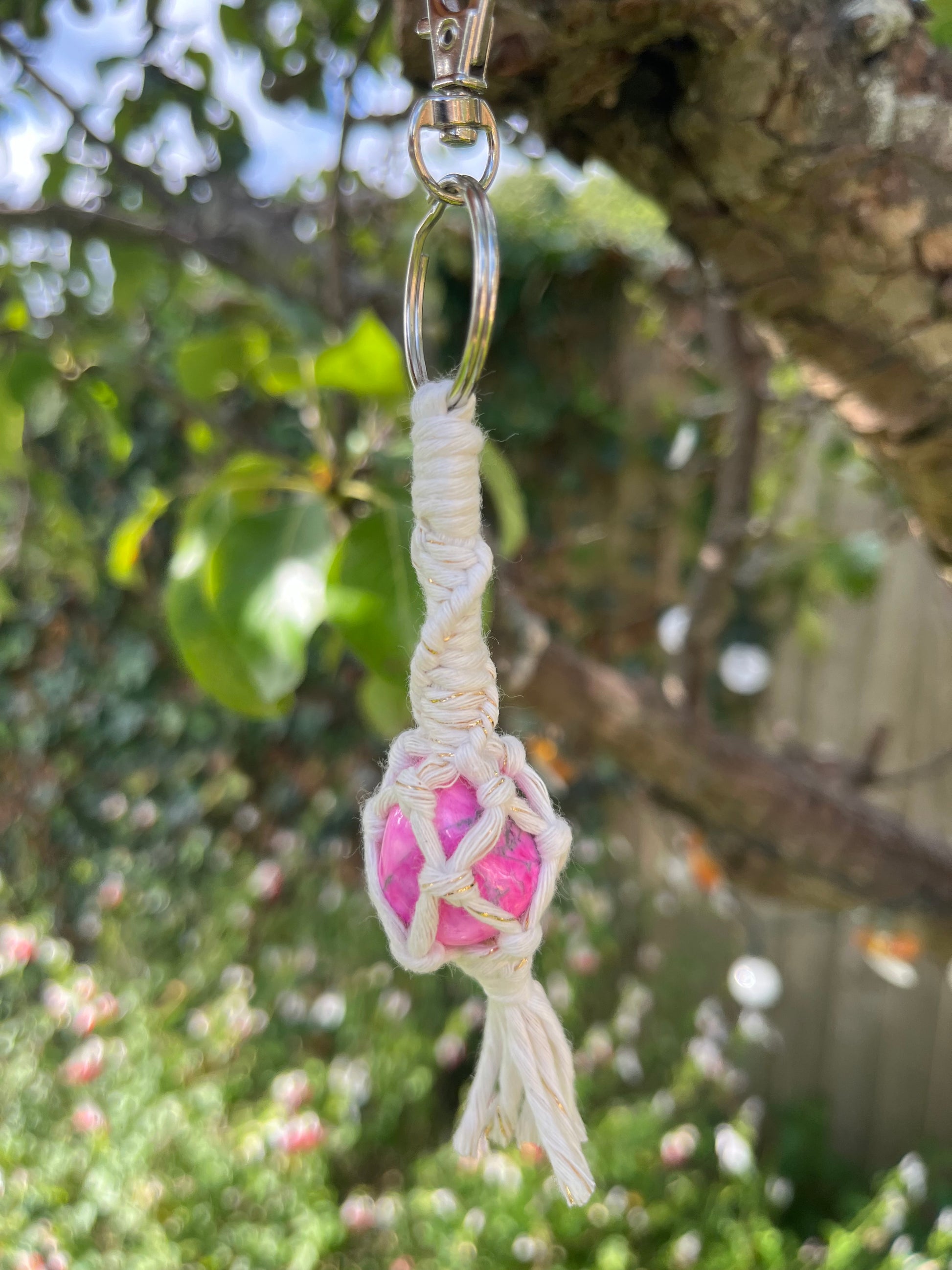 A handmade macrame gemstone keyring, made with natural coloured cotton yarn wrapped around a pink howlite gemstone, and metal split ring clasp, shown hanging outside by a pear tree and other greenery. 