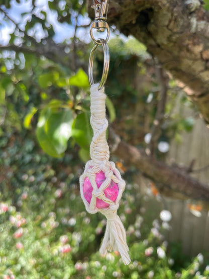 A handmade macrame gemstone keyring, made with natural coloured cotton yarn wrapped around a pink howlite gemstone, and metal split ring clasp, shown hanging outside by a pear tree and other greenery. 