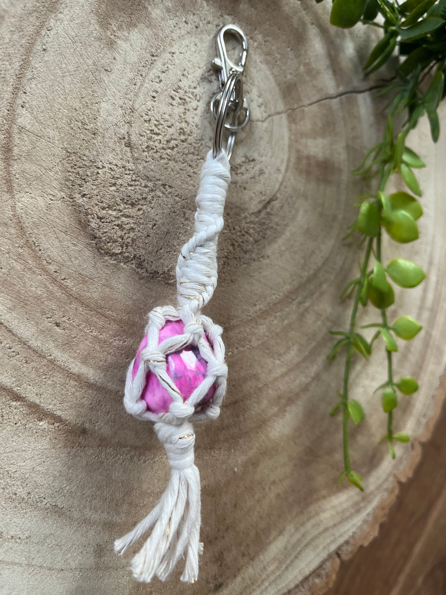 A handmade macrame gemstone keyring, made with natural coloured cotton yarn wrapped around a pink howlite gemstone, and metal split ring clasp, on a wooden surface.