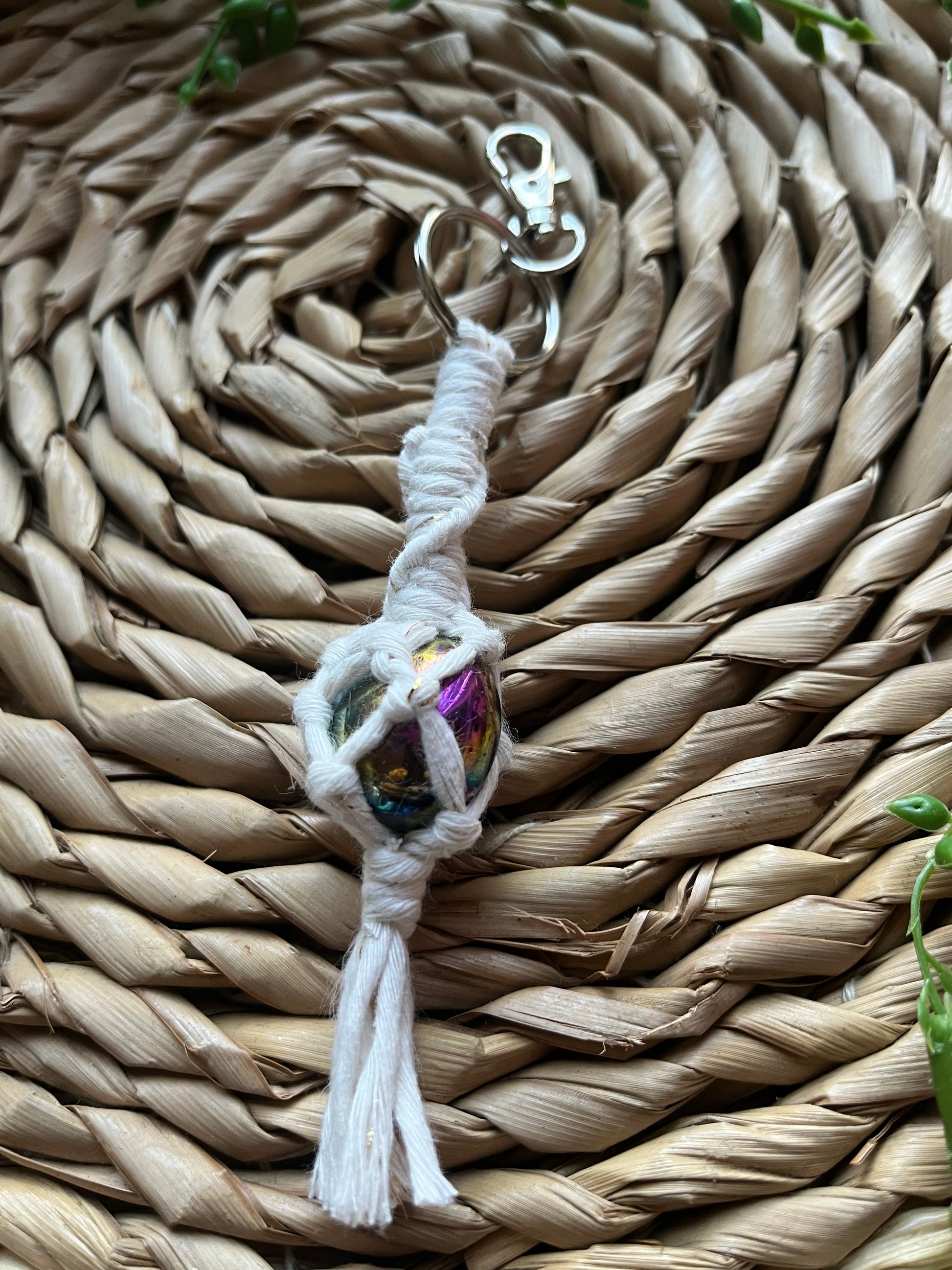 A handmade macrame gemstone keyring, made with natural coloured cotton yarn wrapped around a rainbow aura quartz gemstone, and metal split ring clasp, on a wicker surface.