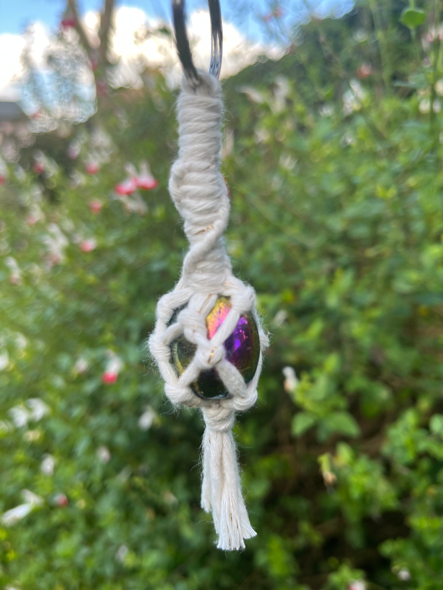 A handmade macrame gemstone keyring, made with natural coloured cotton yarn wrapped around a rainbow aura quartz gemstone, displayed hanging outside with lush green plants in the background. 