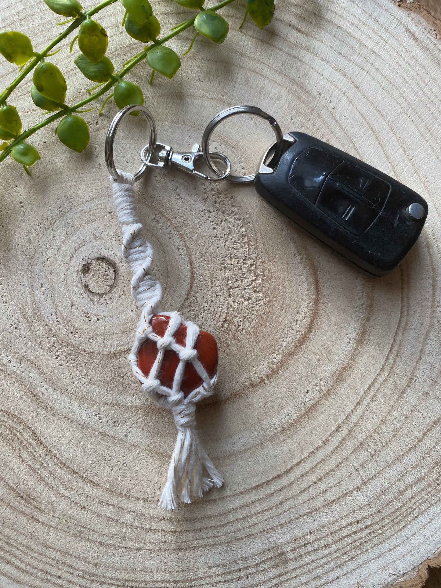 A handmade macrame gemstone keyring, made with natural coloured cotton yarn wrapped around a res jasper gemstone, and metal split ring clasp, on a wooden surface attached to a single car key. 