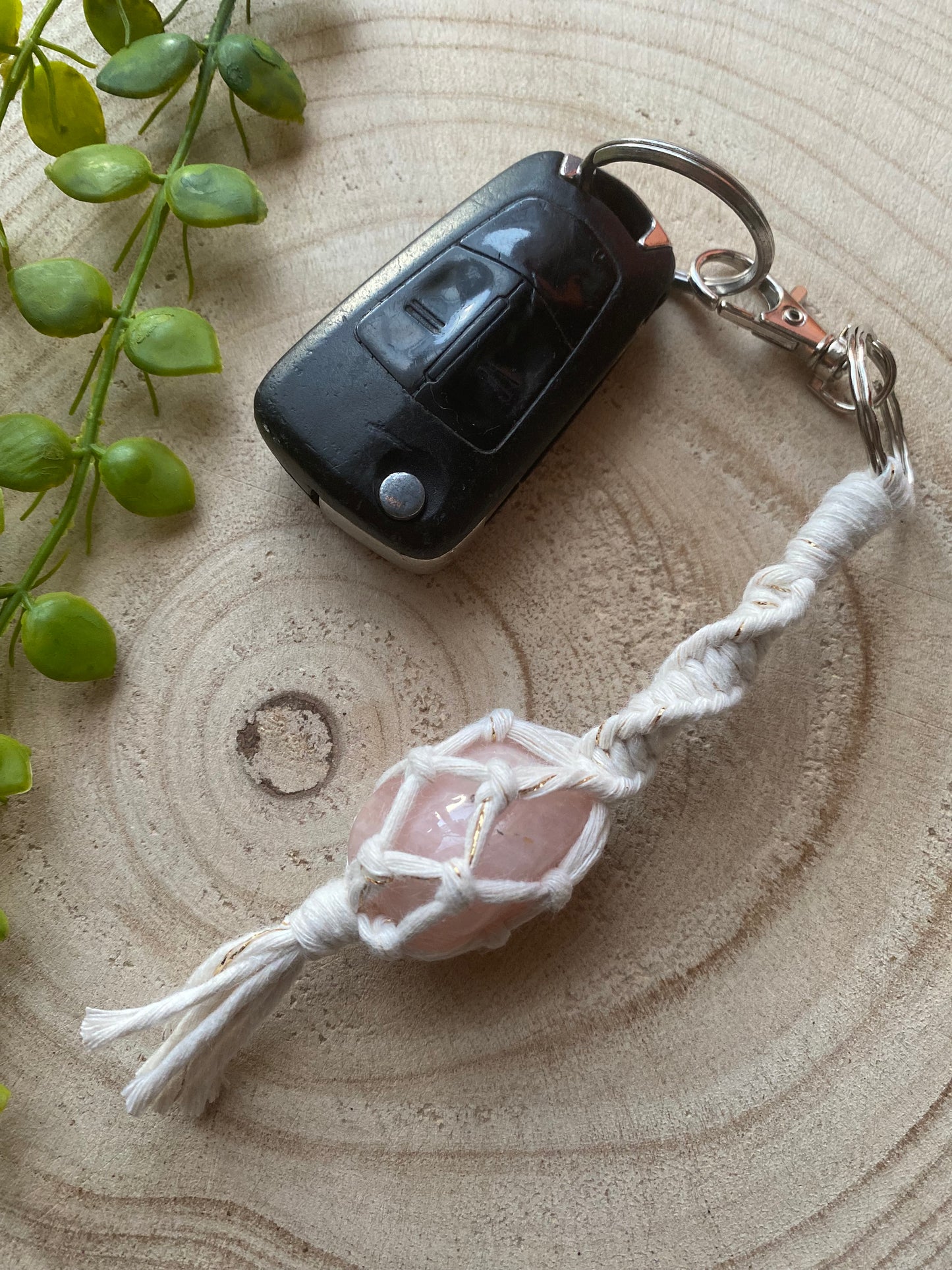 A handmade macrame gemstone keyring, made with natural coloured cotton yarn wrapped around a pink rose quartz gemstone, and metal split ring clasp, on a wooden surface attached to a single car key. 