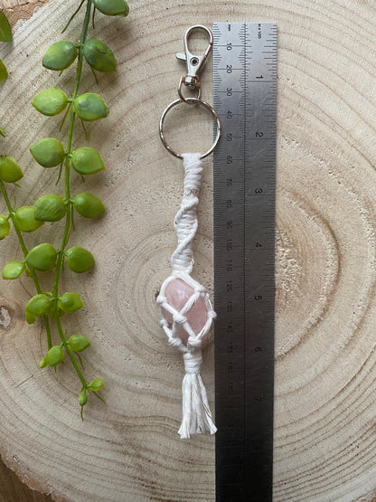 A handmade macrame gemstone keyring, made with natural coloured cotton yarn wrapped around a pink rose quartz gemstone, and metal split ring clasp, on a wooden surface next to a metal rule. 