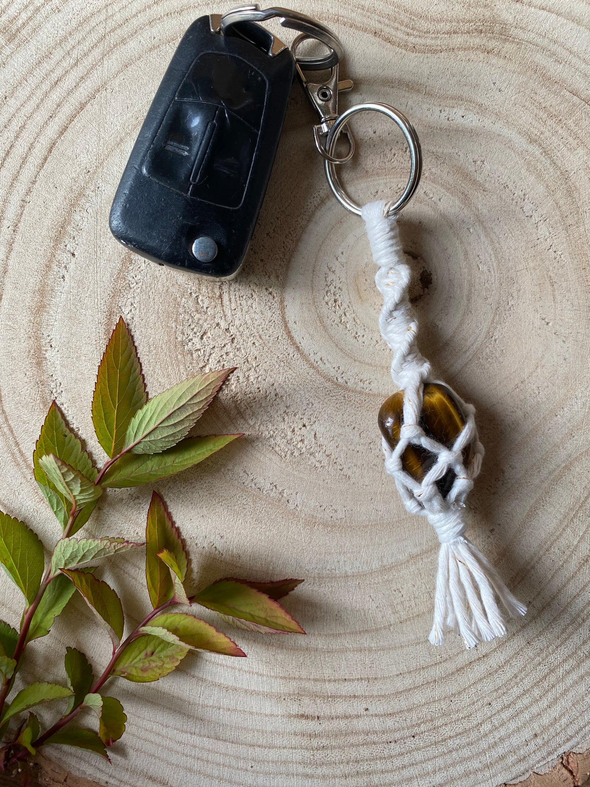 A handmade macrame gemstone keyring, made with natural coloured cotton yarn wrapped around a golden tigers eye gemstone, and metal split ring clasp, on a wooden surface attached to a single car key. 