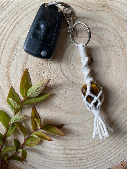 A handmade macrame gemstone keyring, made with natural coloured cotton yarn wrapped around a golden tigers eye gemstone, and metal split ring clasp, on a wooden surface attached to a single car key. 