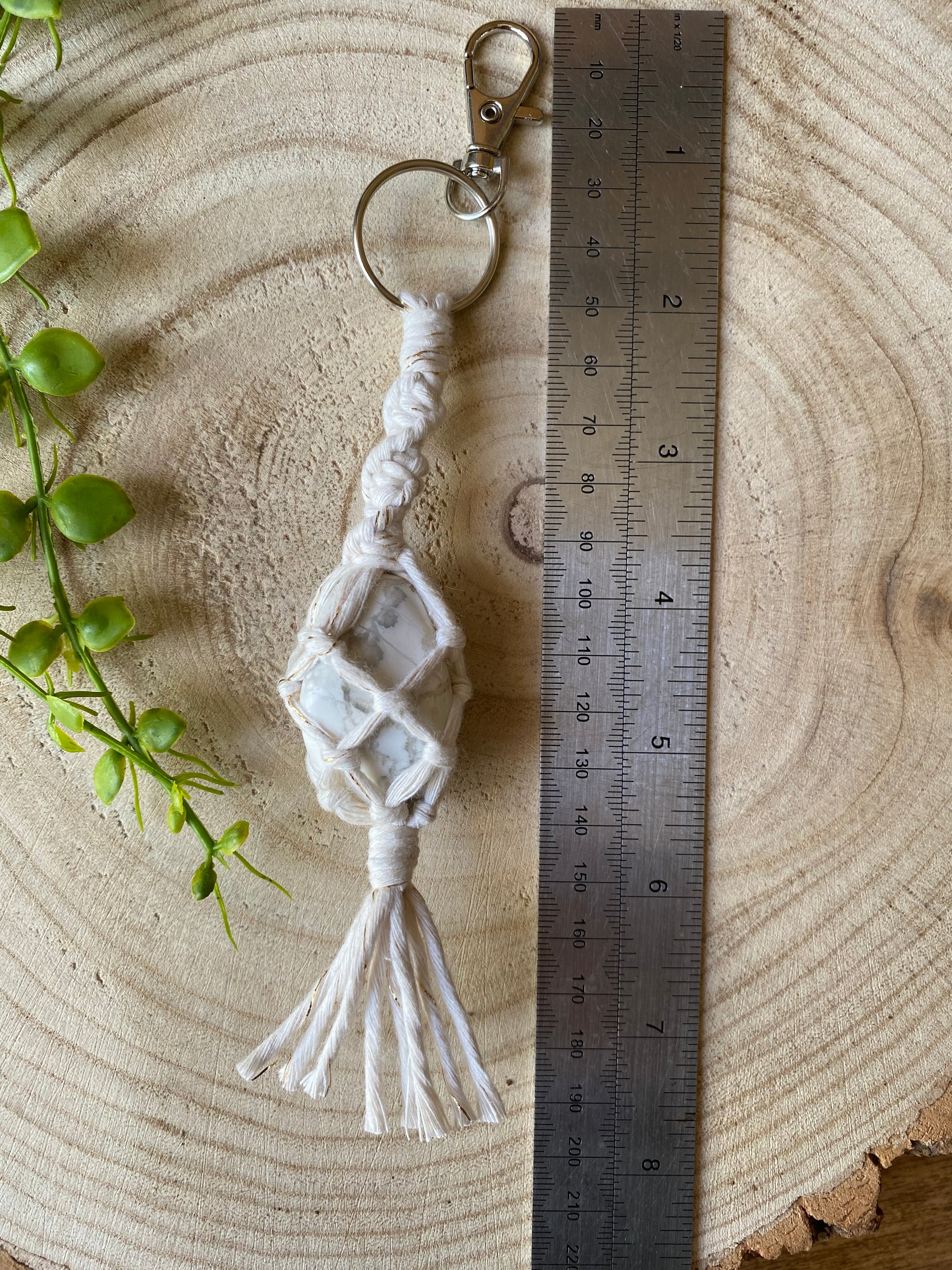 A handmade macrame gemstone keyring, made with natural coloured cotton yarn wrapped around a white howlite gemstone, and metal split ring clasp, on a wooden surface next to a metal rule. 