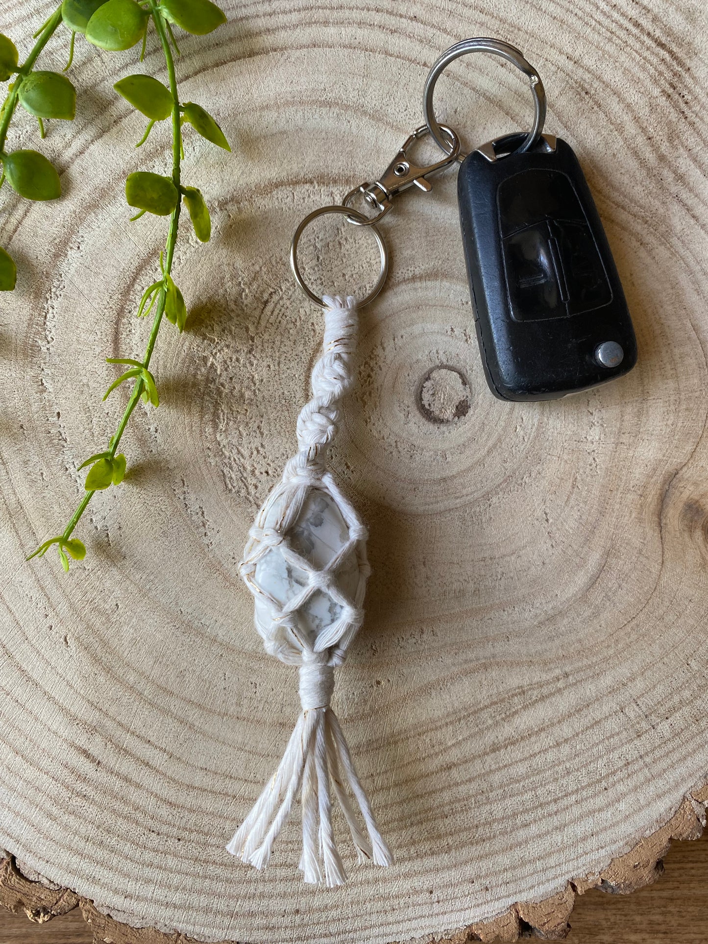 A handmade macrame gemstone keyring, made with natural coloured cotton yarn wrapped around a white howlite gemstone, and metal split ring clasp, on a wooden surface attached to a single car key. 