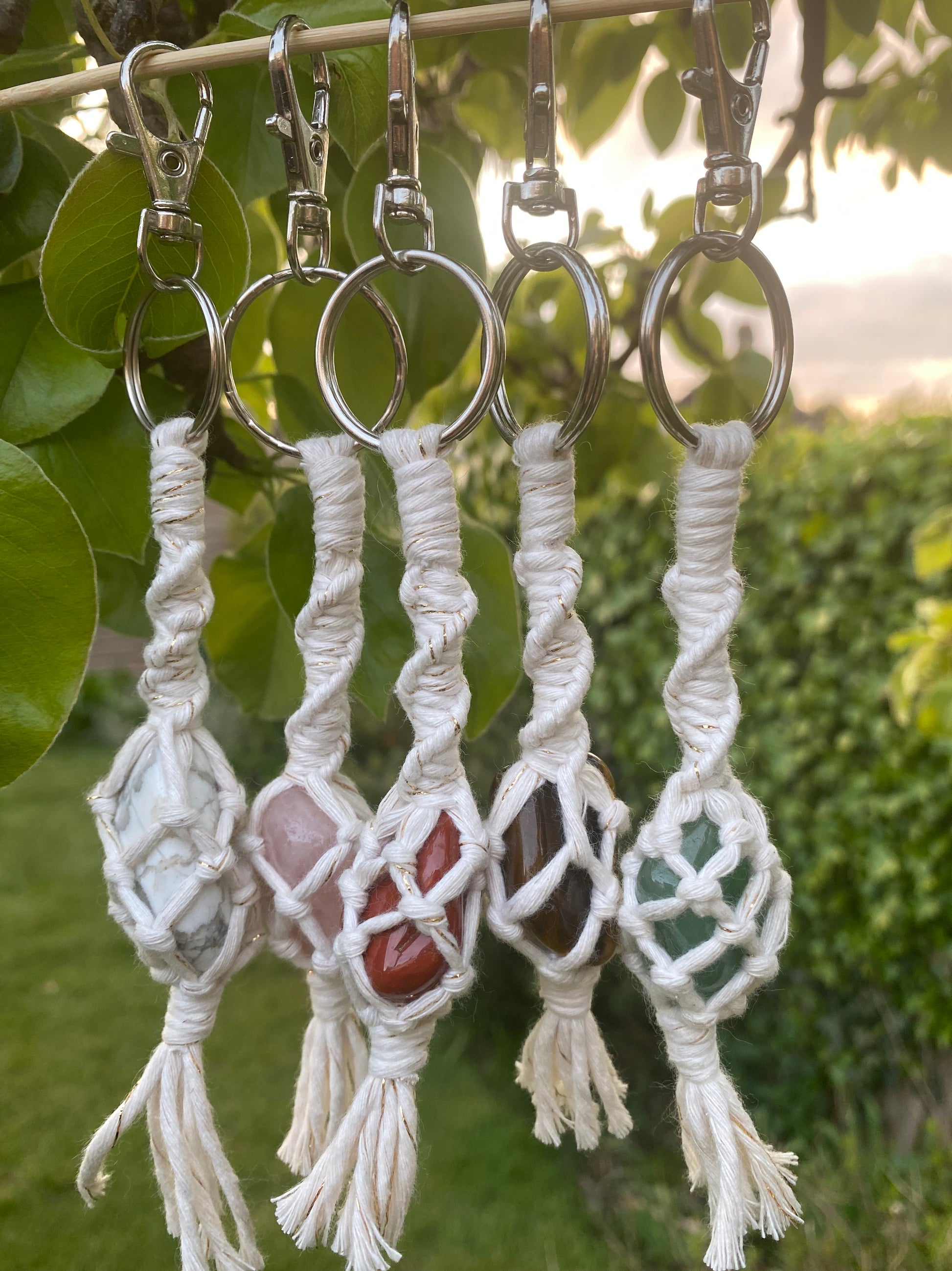 5 handmade macrame gemstone keyrings, made with golden natural cotton yarn wrapped around a variety of gemstones, with metal split ring clasps showing hanging outside with lush greenery surrounding them. 