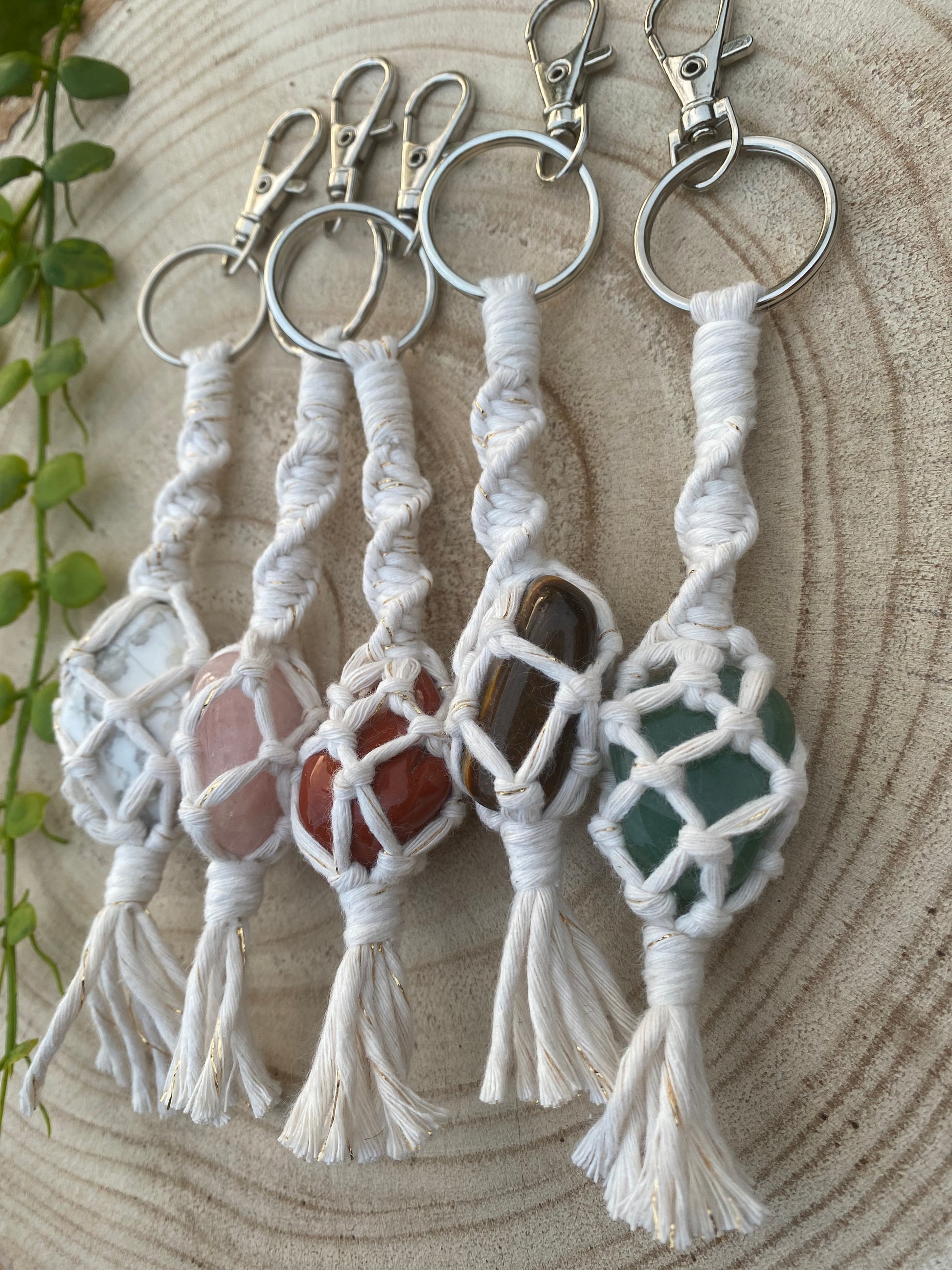 A selection of handmade macrame gemstone keyrings, made with natural coloured cotton yarn wrapped around different coloured gemstones, laid out on a wooden surface next to each other.