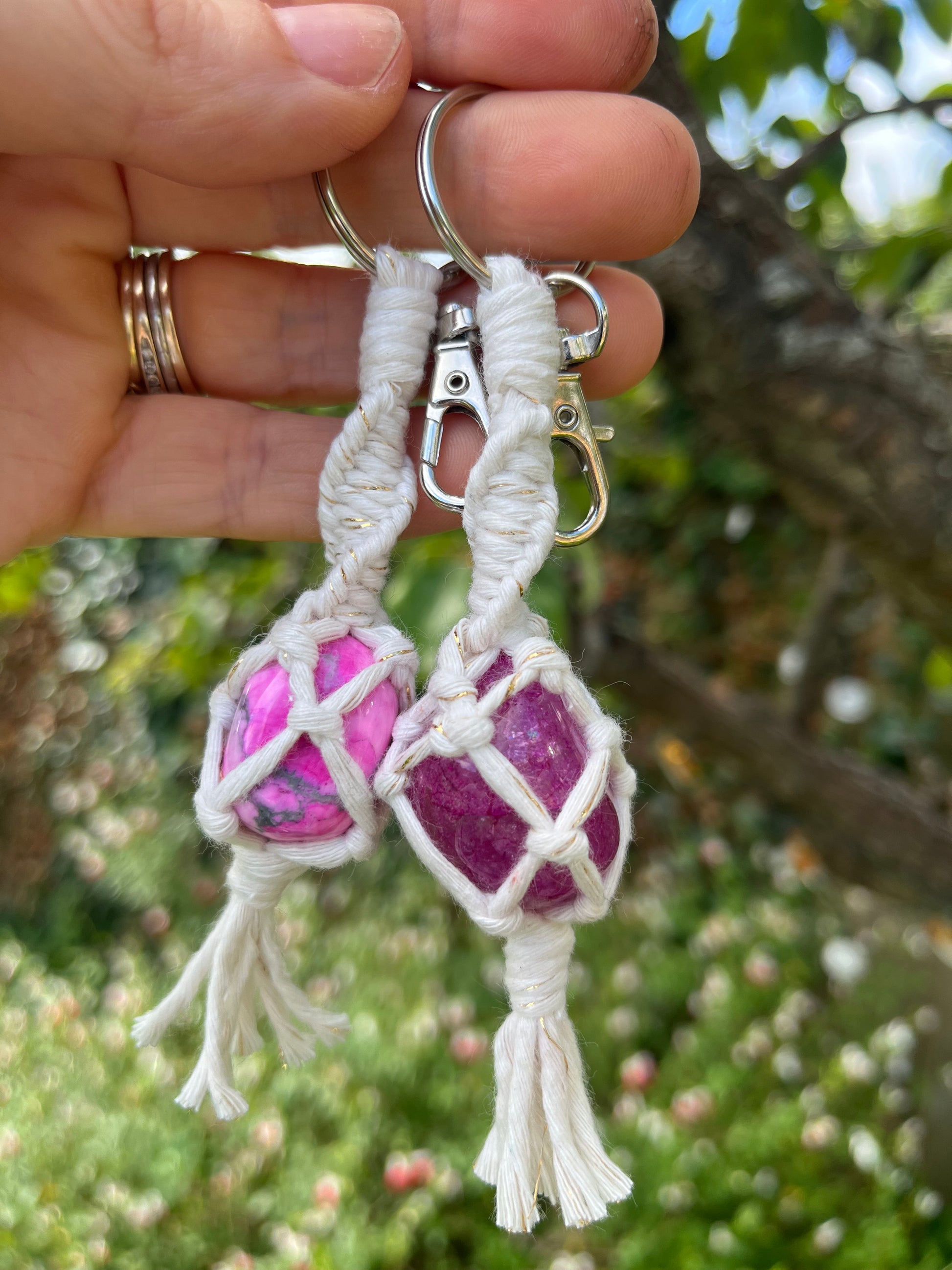 A hand holds two handmade macrame gemstone keyrings, made with natural coloured cotton yarn wrapped around pink gemstones. The background shows out of focus greenery. 