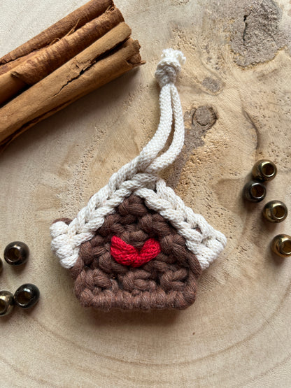 A small macramé decoration in the shape of a gingerbread house, made with brown and white yarn with a red heart detail in the middle, styled on a light wood surface with cinnamon sticks and metal beads. 