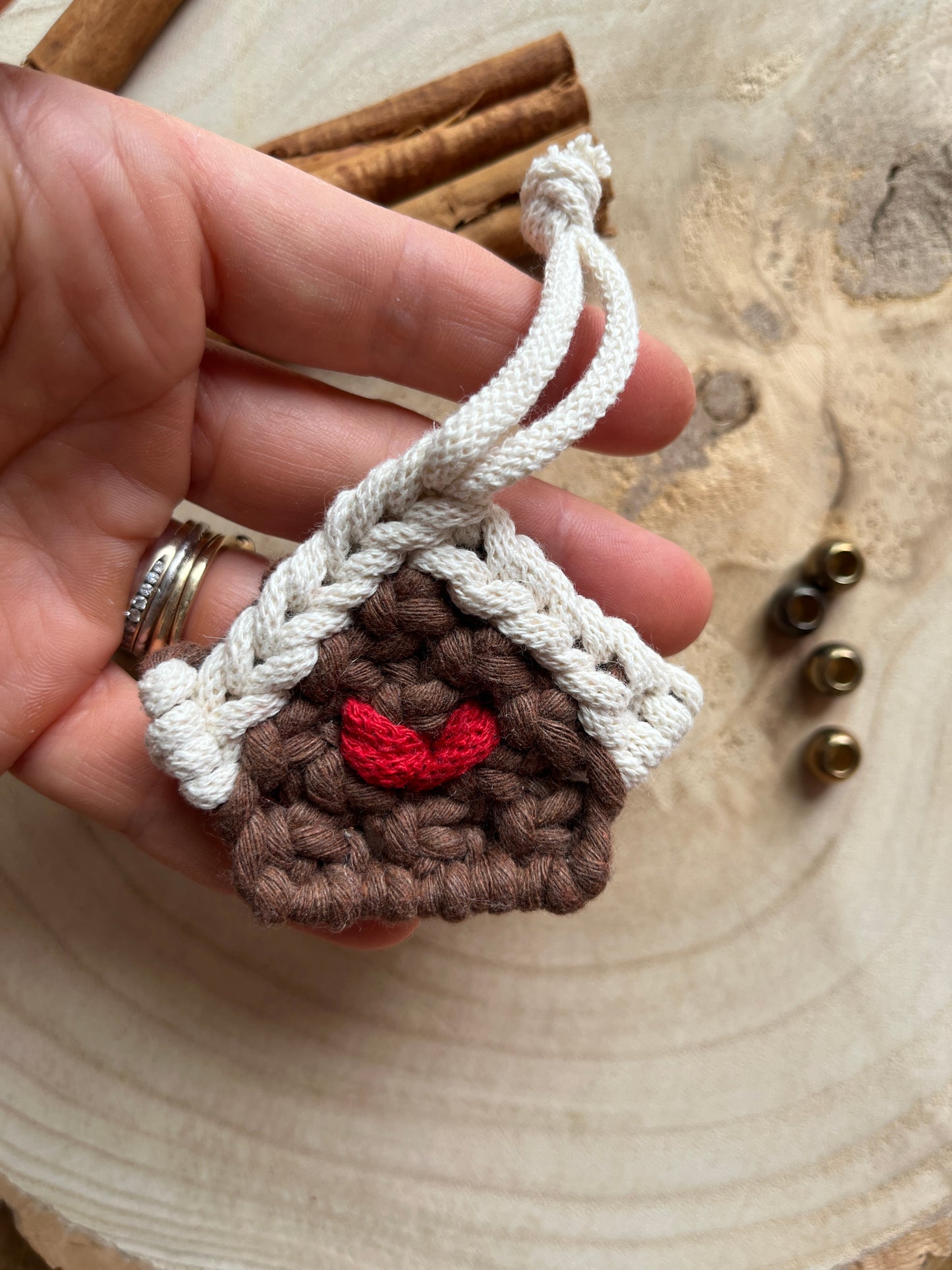 A hand holding a small macramé decoration in the shape of a gingerbread house, made with brown and white yarn with a red heart detail in the middle. 