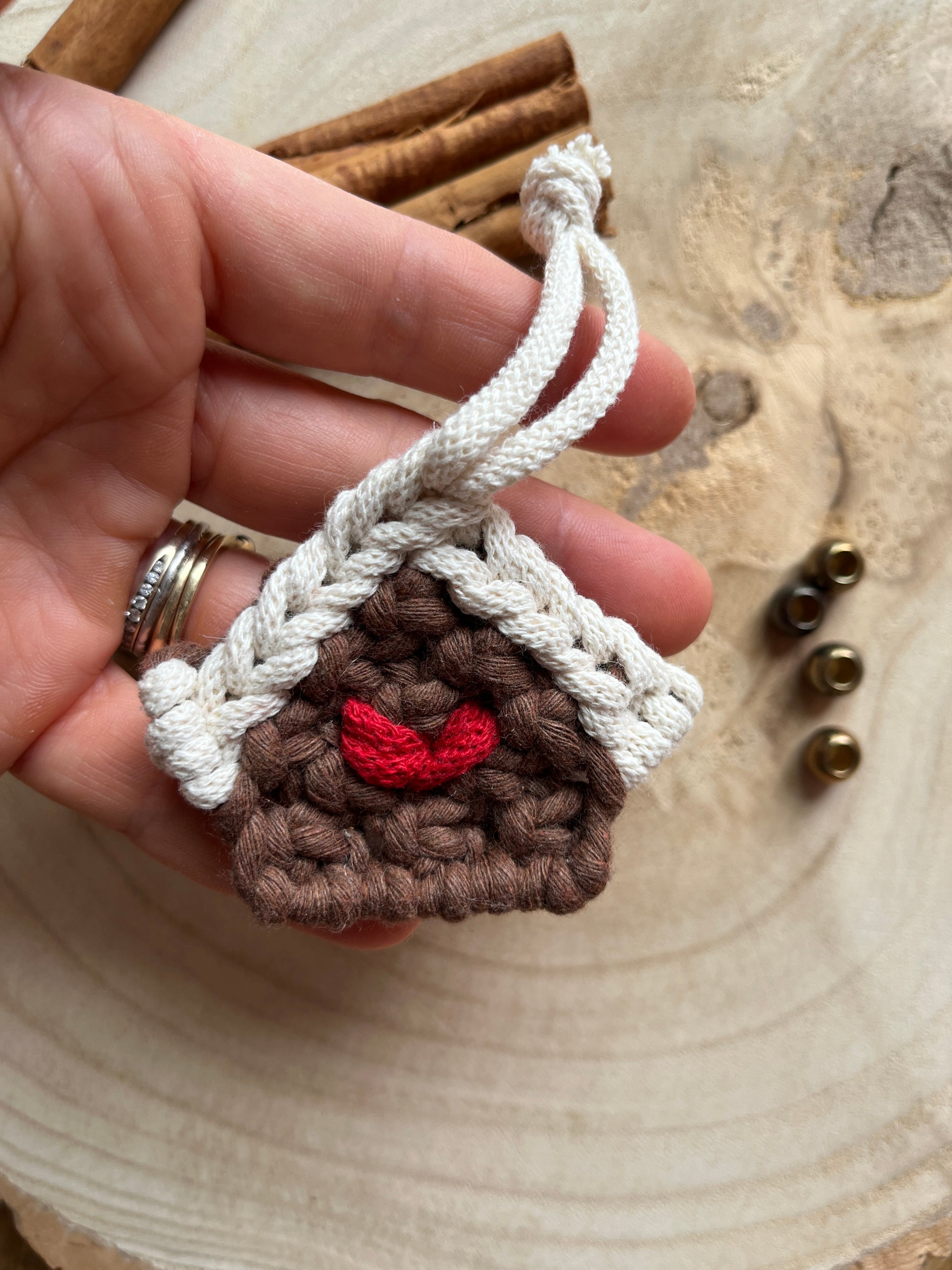 A hand holding a small macramé decoration in the shape of a gingerbread house, made with brown and white yarn with a red heart detail in the middle. 
