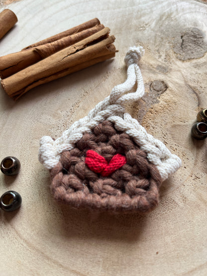 Close up of a small macramé decoration in the shape of a gingerbread house, made with brown and white yarn with a red heart detail in the middle, styled on a light wood surface with cinnamon sticks and metal beads. 