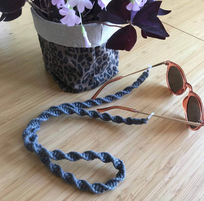 Sunglasses with a dark grey strap on a wooden surface next to a leopard print pot with flowers.