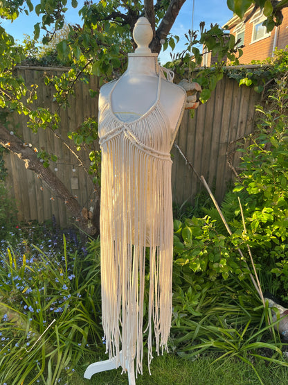 A beige, one-size macrame boho goddess dress with long fringe on a mannequin outdoors.
