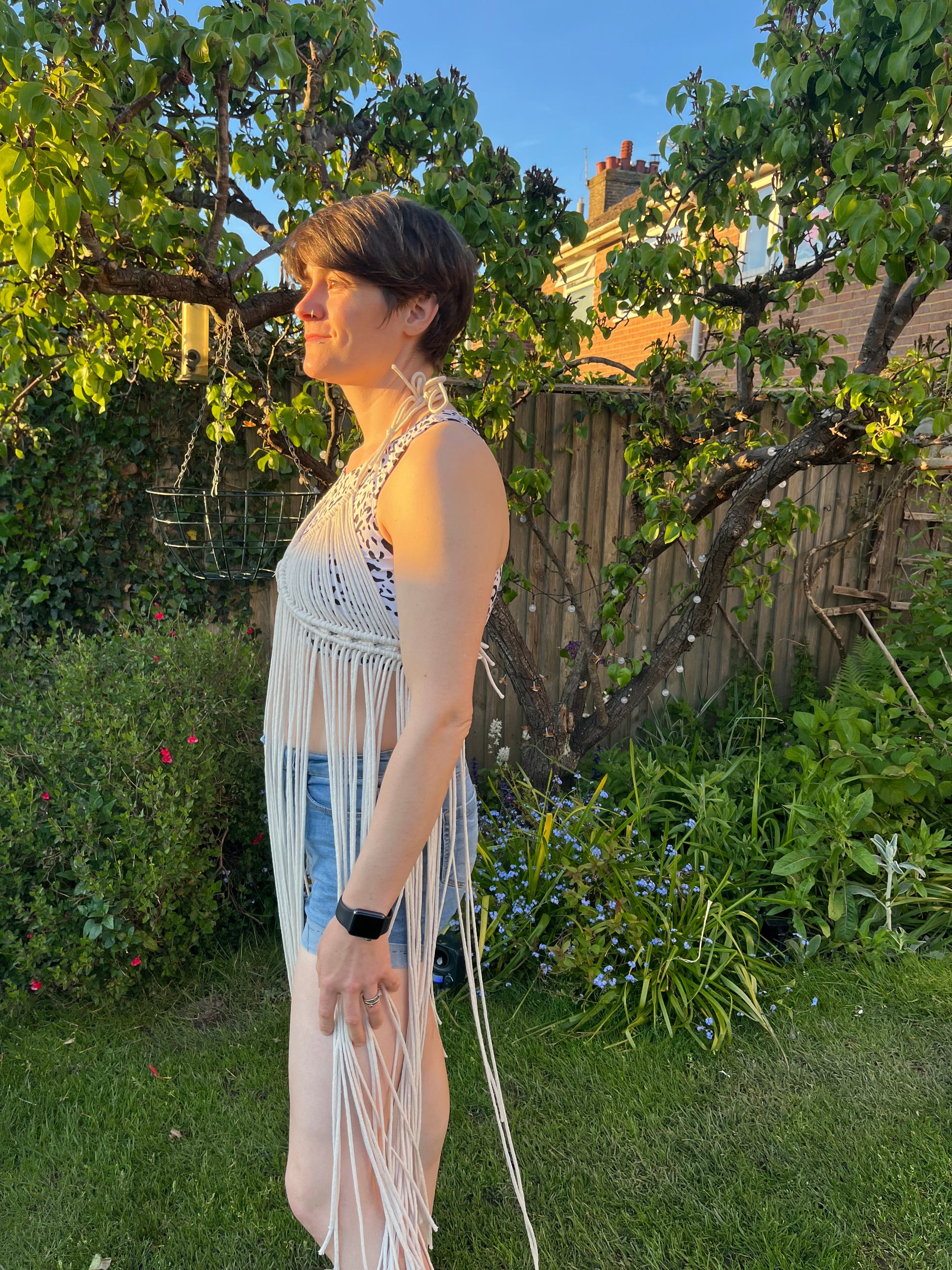 A woman with short brown hair stands outside wearing a natural macrame boho goddess dress coverup with halter neck over denim shorts, she is turned sideways to the camera. 