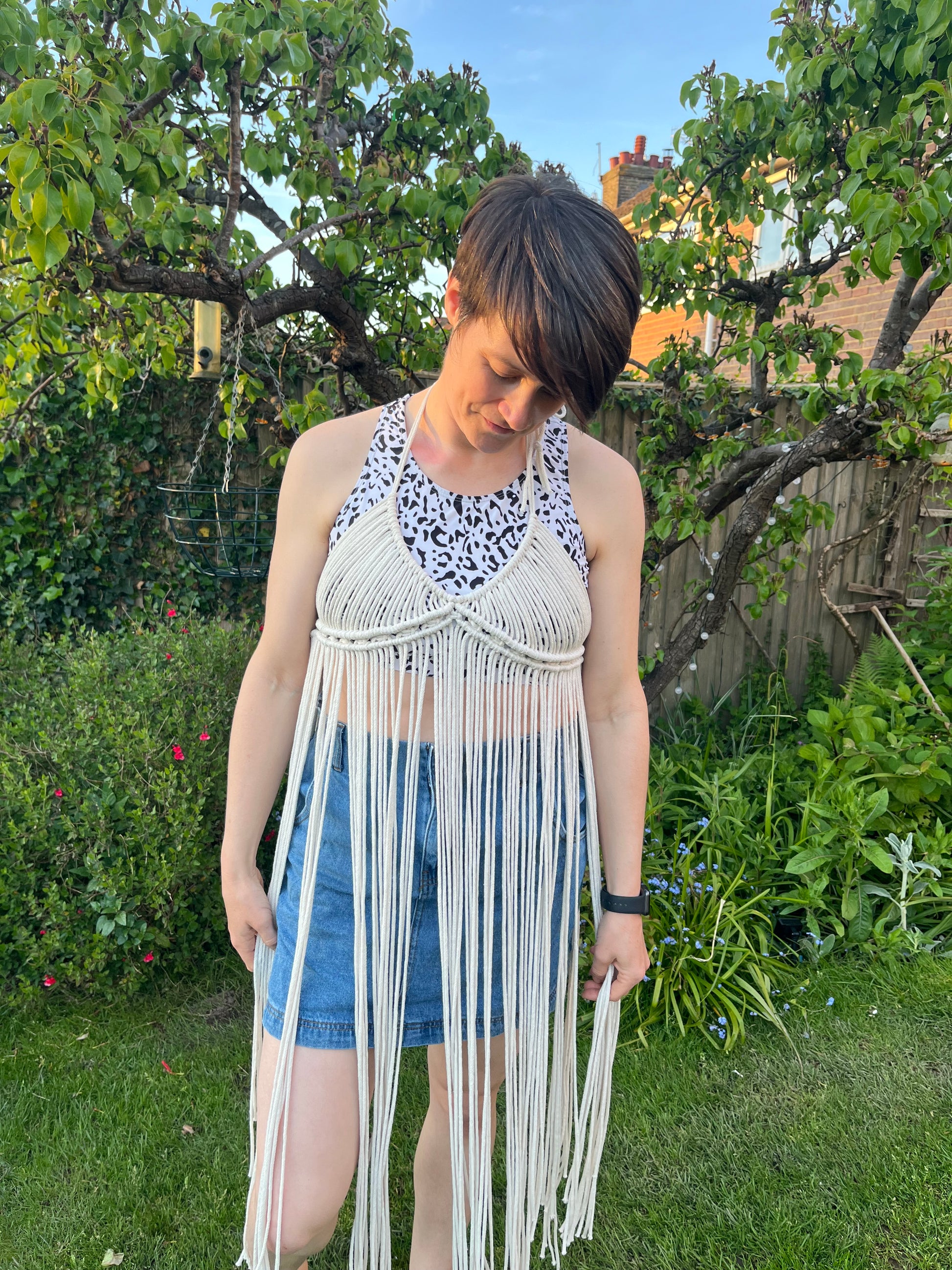 A woman with short brown hair stands outside wearing a natural macrame boho goddess dress coverup with halter neck over a patterned crop top and denim shorts, she is looking down to the floor and smiling. 