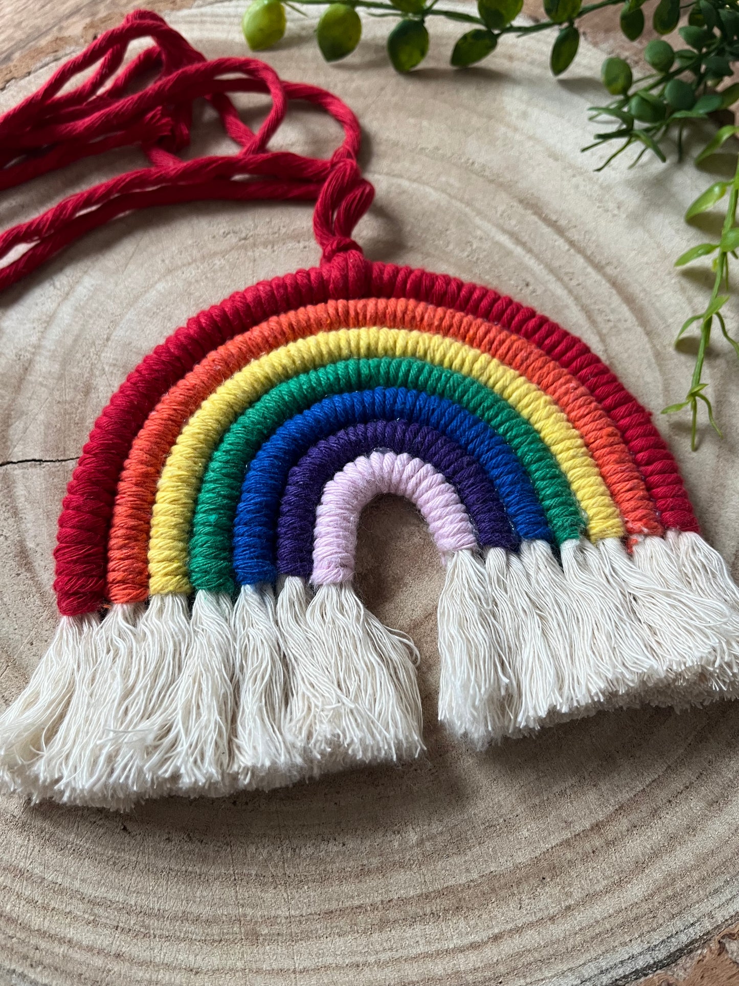 Close up of a handmade macrame rainbow wall hanging with vibrant colours, made from recycled cotton yarn, with a red hanging cord and white tassels at the bottom, on a wooden board. 