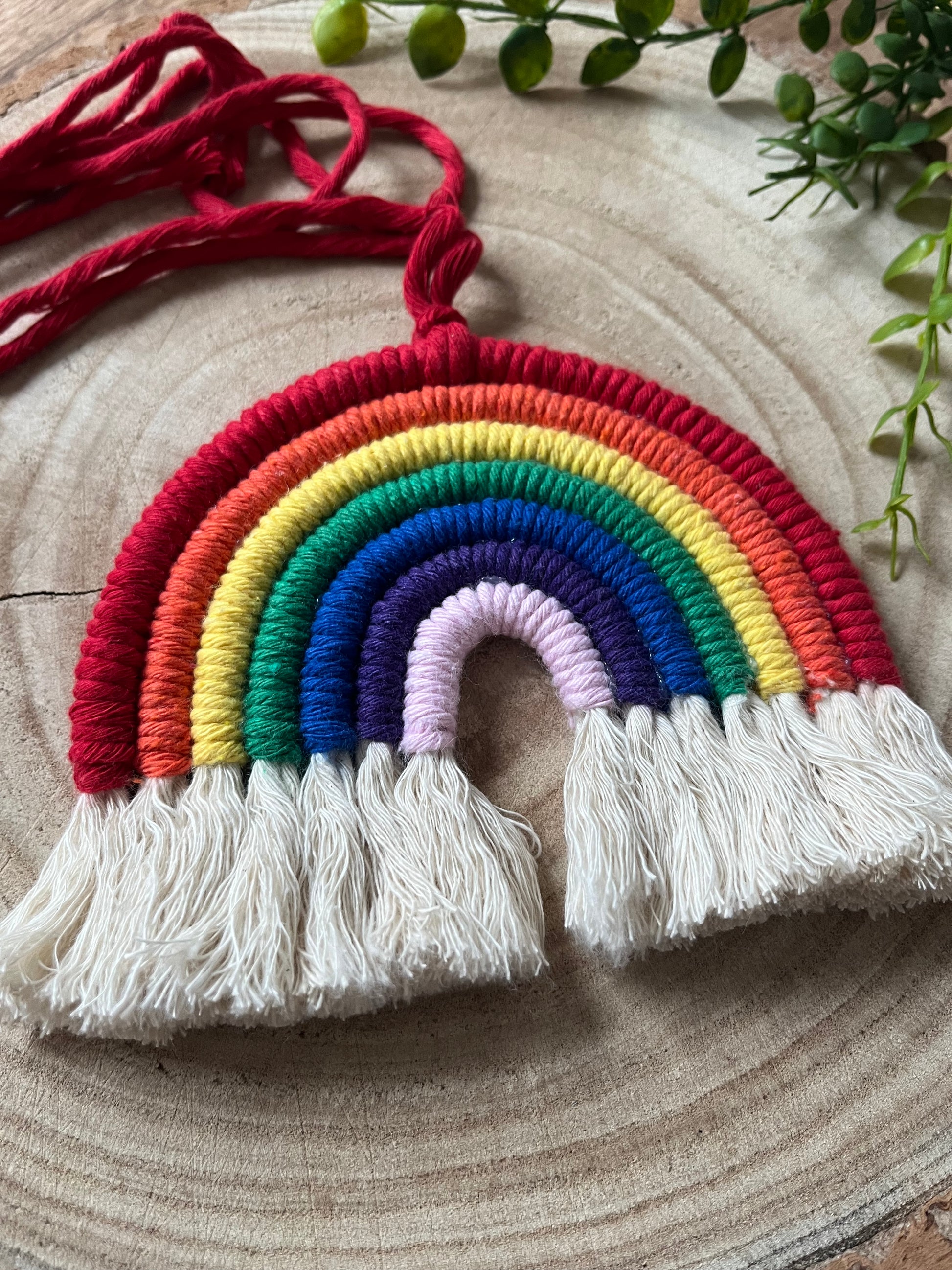 Close up of a handmade macrame rainbow wall hanging with vibrant colours, made from recycled cotton yarn, with a red hanging cord and white tassels at the bottom, on a wooden board. 