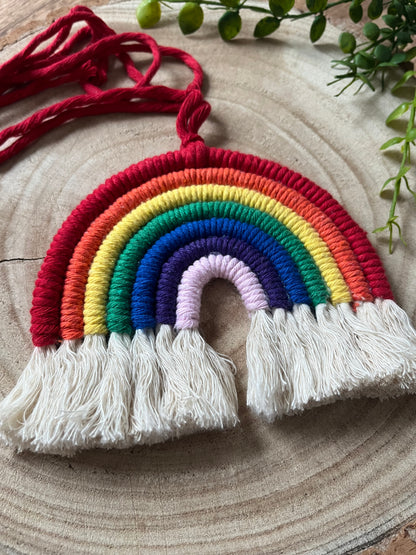 Close up of a handmade macrame rainbow wall hanging with vibrant colours, made from recycled cotton yarn, with a red hanging cord and white tassels at the bottom, on a wooden board. 