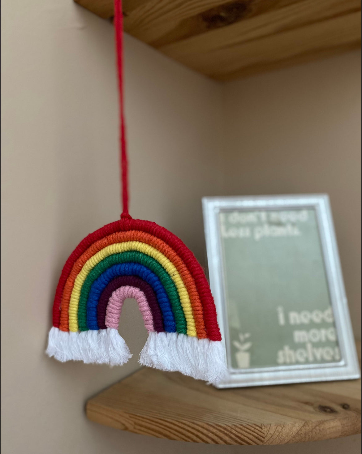 A handmade macrame rainbow wall hanging with vibrant colours, made from recycled cotton yarn, with a red hanging cord and white tassels at the bottom hanging from a wooden corner shelf next to a framed print. 