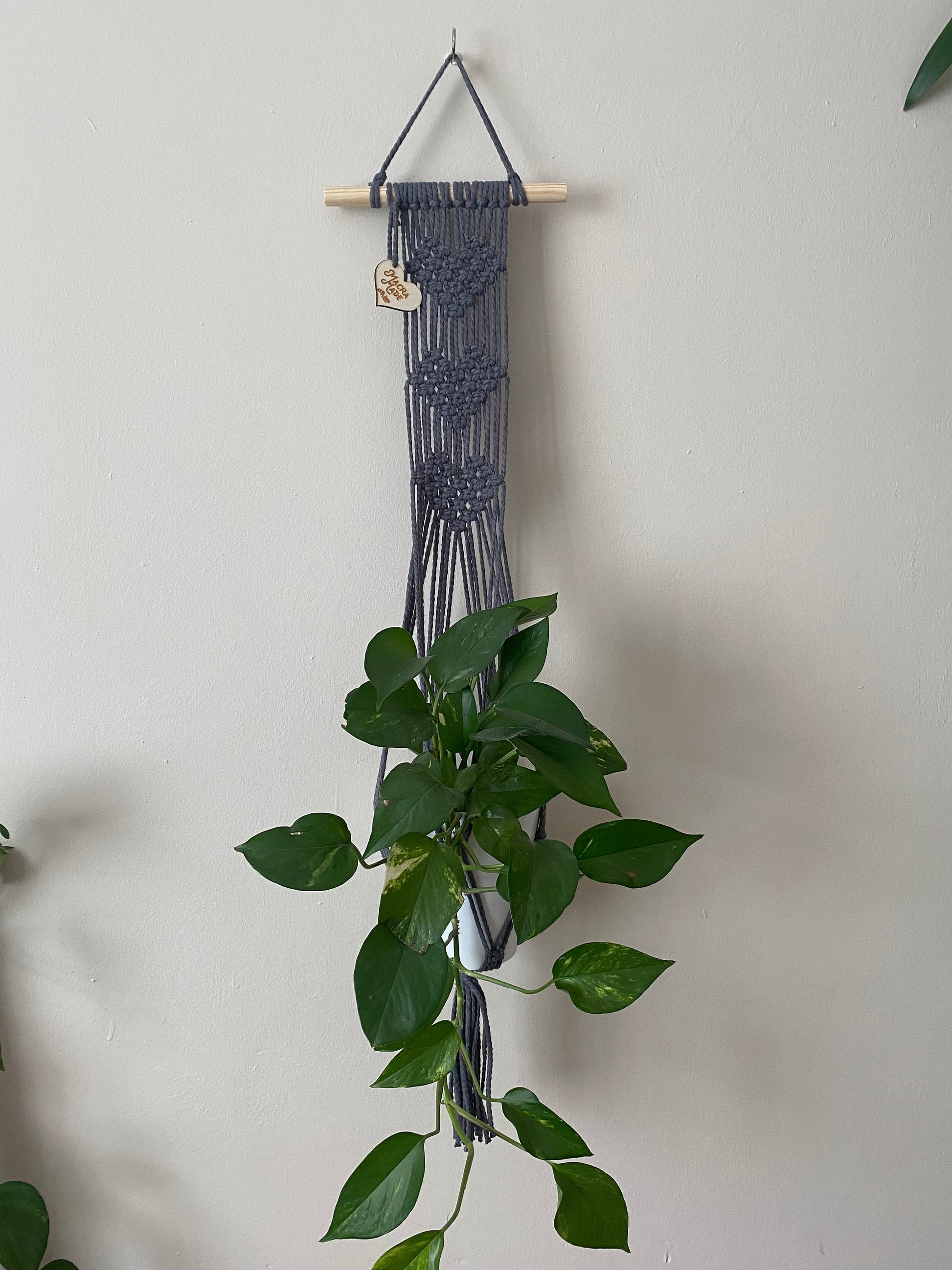 A trailing green plant in a ceramic pot, sits inside a long macrame wall hanging plant hanger with heart detail, made with slate grey yarn on a small wooden dowel. 
