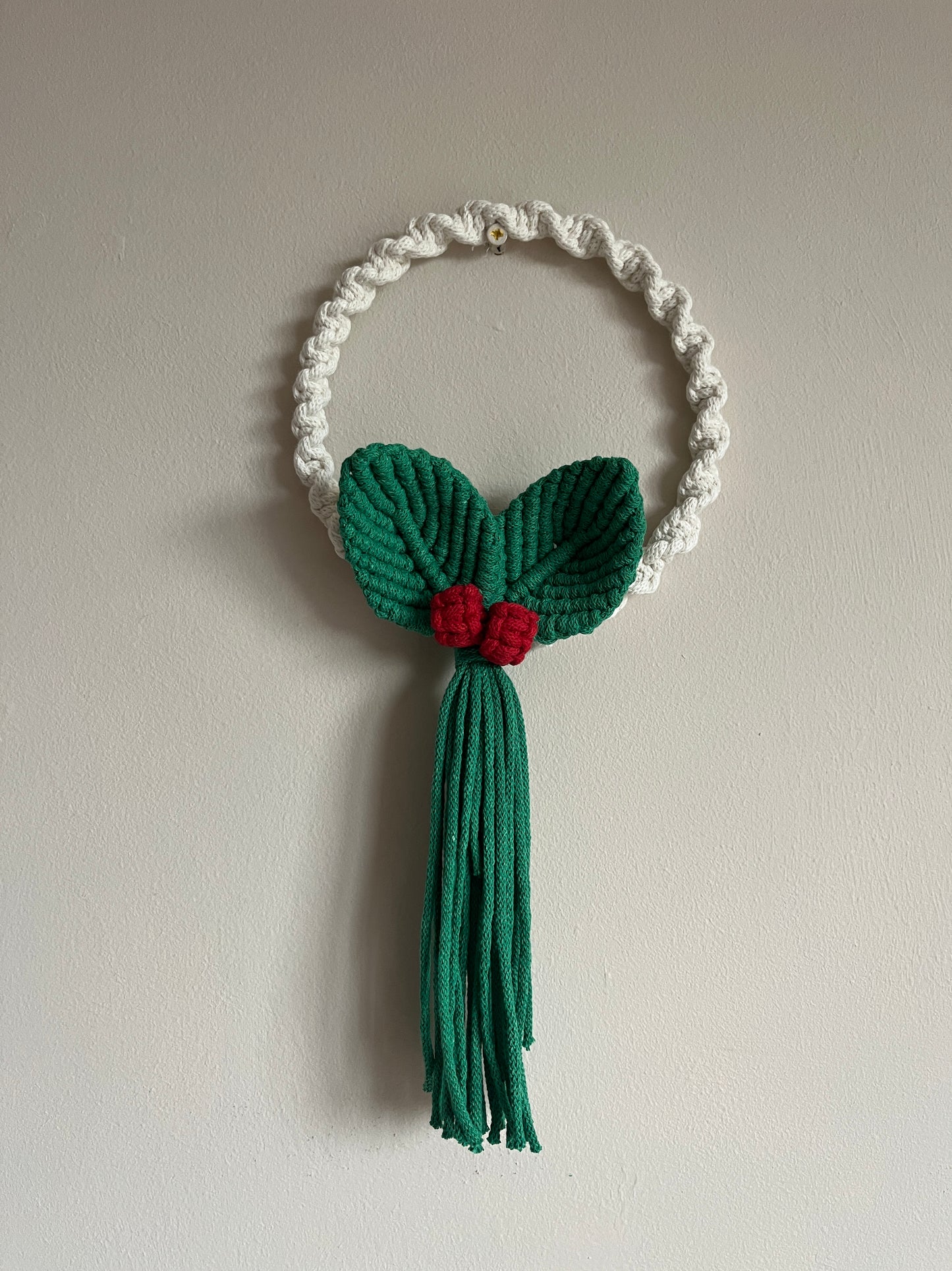 White knotted circular wall hanging macrame wreath with green leaves and red berries on a beige wall