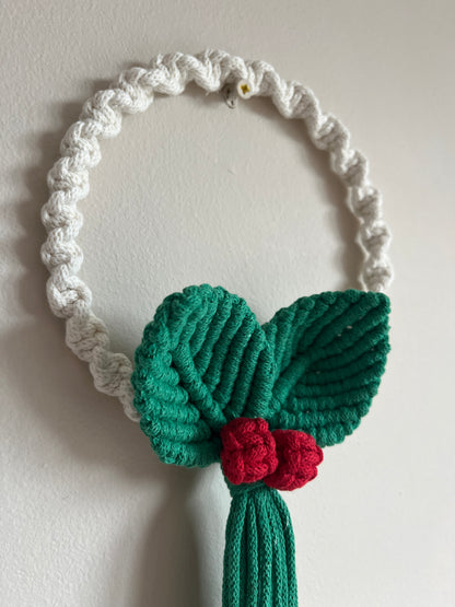 Close up of a white knotted circular wall hanging macrame wreath with green leaves and red berries on a beige wall