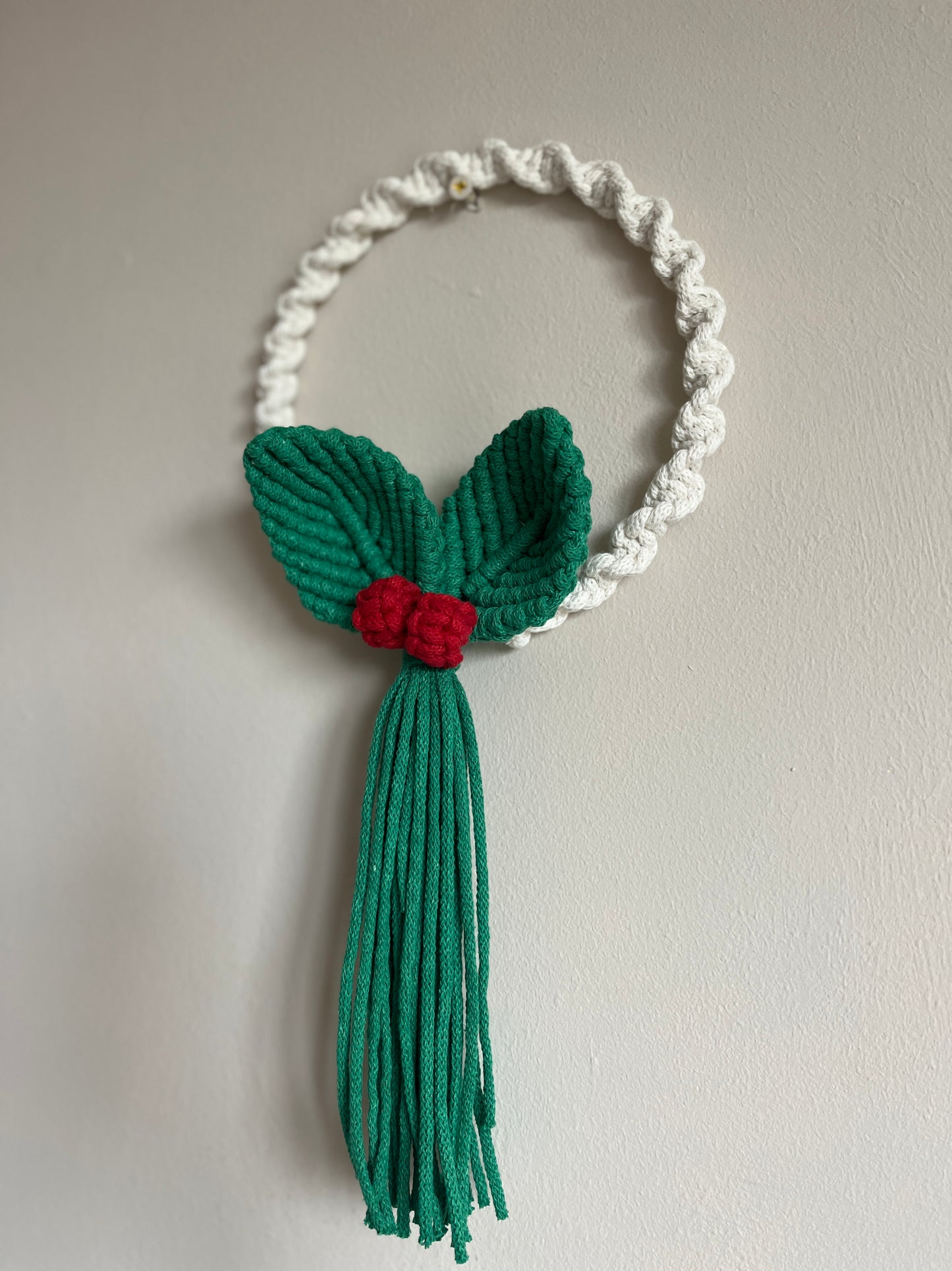 White knotted handmade wall hanging macrame wreath with green leaves and red berries with long green tails, on a beige wall