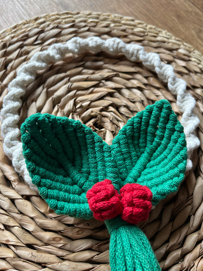 Handmade circular wall hanging macrame wreath with green leaves and red berries, placed on a woven mat