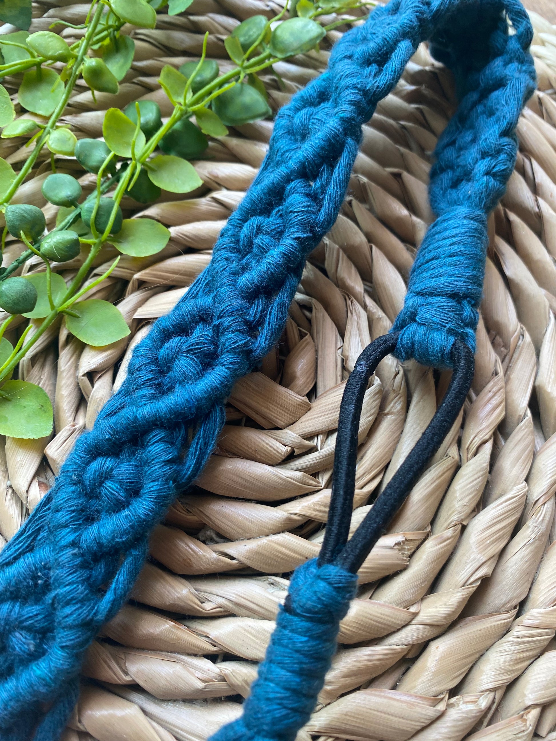 Close up of a blue knotted headband made from recycled cotton cord with an elastic finish, displayed on a woven placemat with green plant vines.
