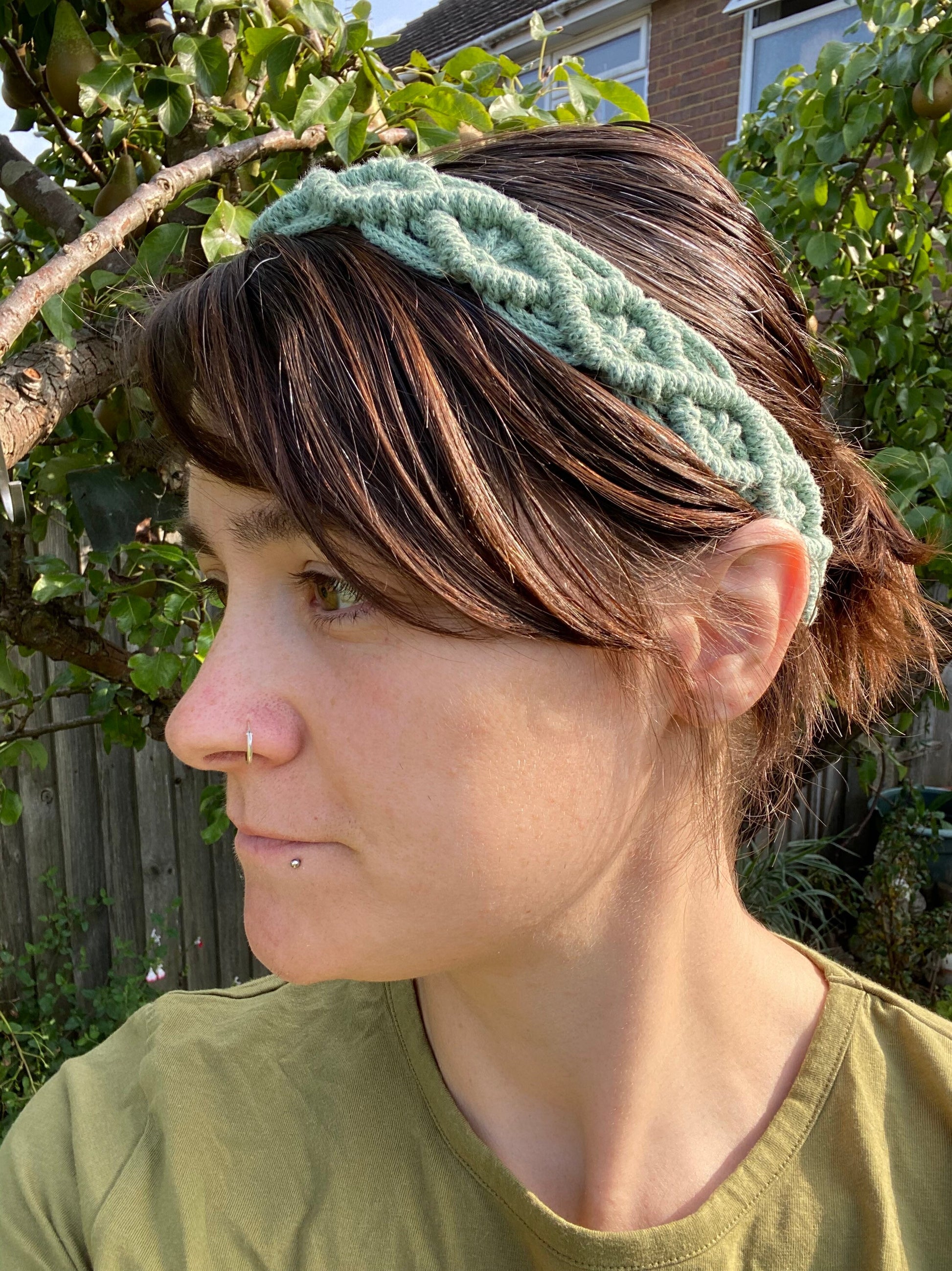 A woman with long brown hair stands outside wearing a green knotted headband made from recycled cotton cord with an infinite knot pattern. 
