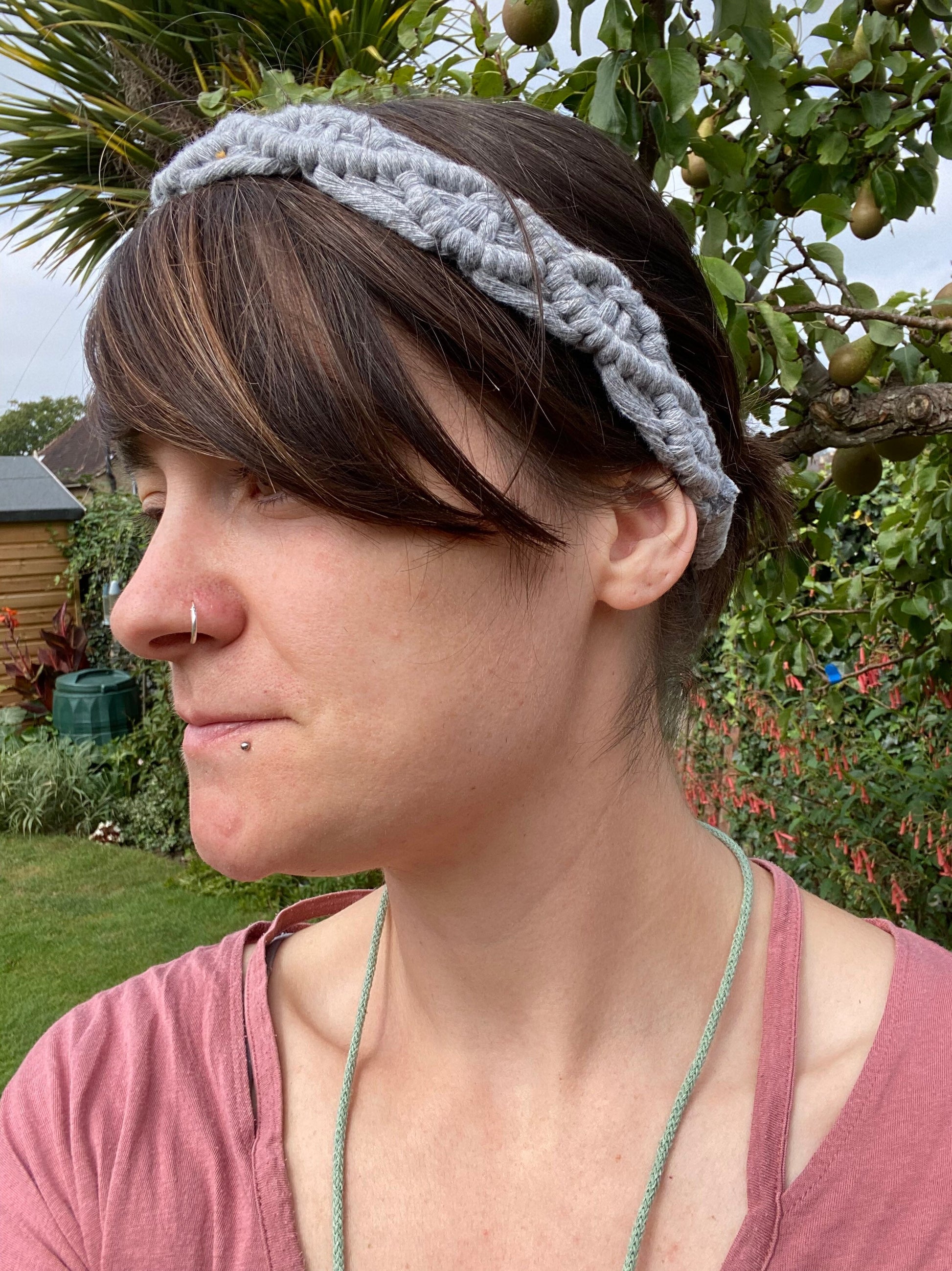 A woman with long brown hair stands outside wearing a grey knotted headband made from recycled cotton cord with an infinite knot pattern. 