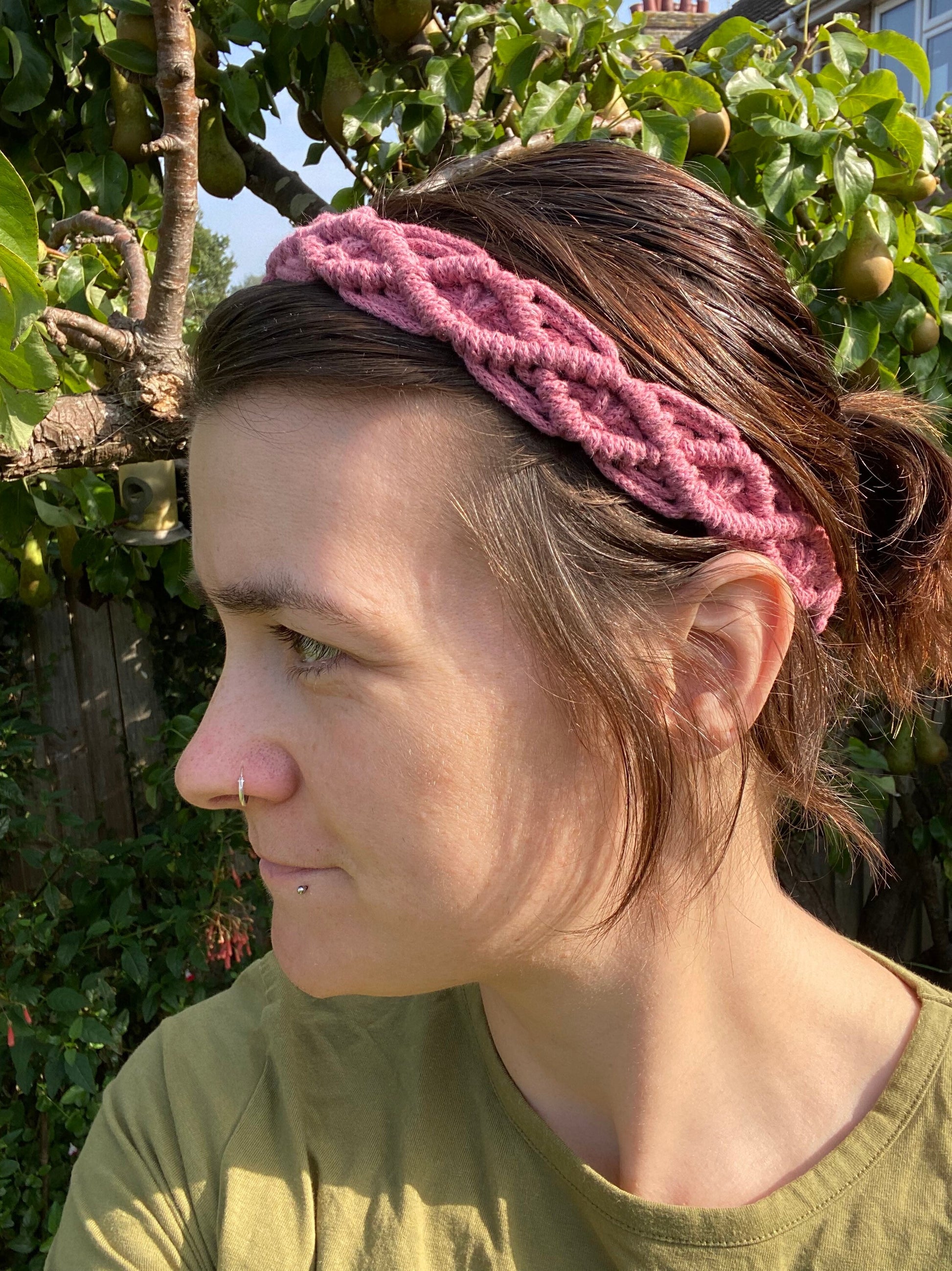 A woman with long brown hair stands outside wearing a pink knotted headband made from recycled cotton cord with an infinite knot pattern. 