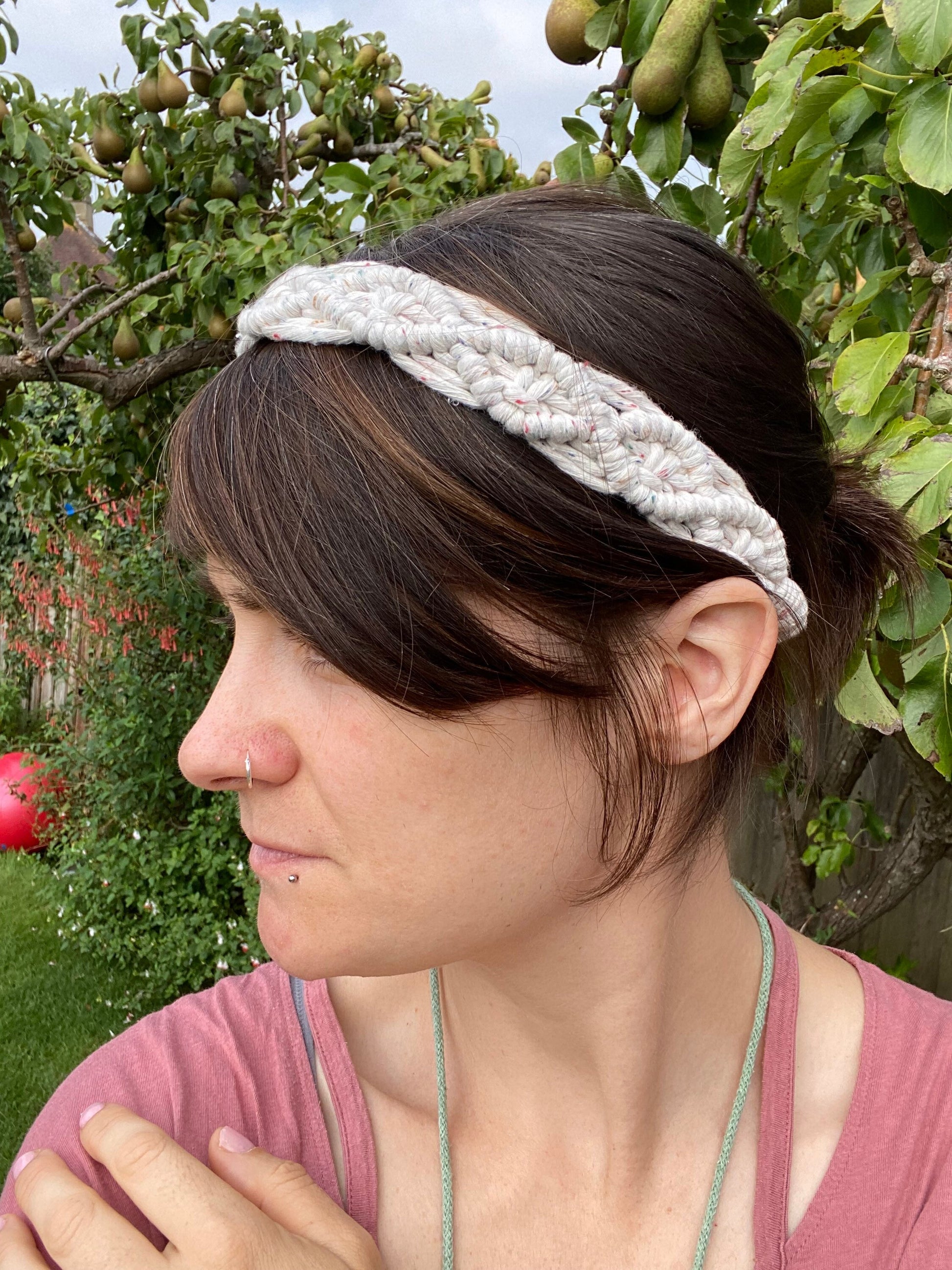 A woman with long brown hair stands outside wearing a white knotted headband made from recycled cotton cord with an infinite knot pattern. 