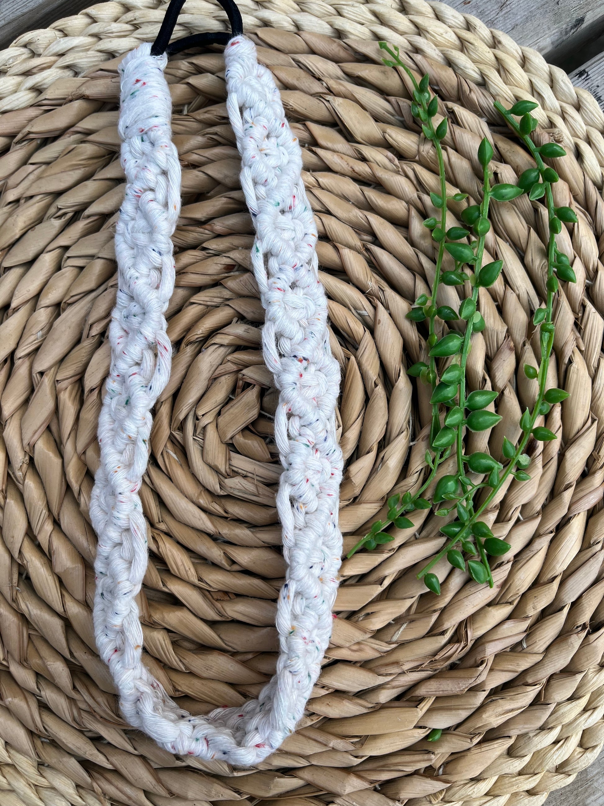 A knotted headband made from recycled cotton cord with an elastic finish, displayed on a woven placemat with green plant vines.