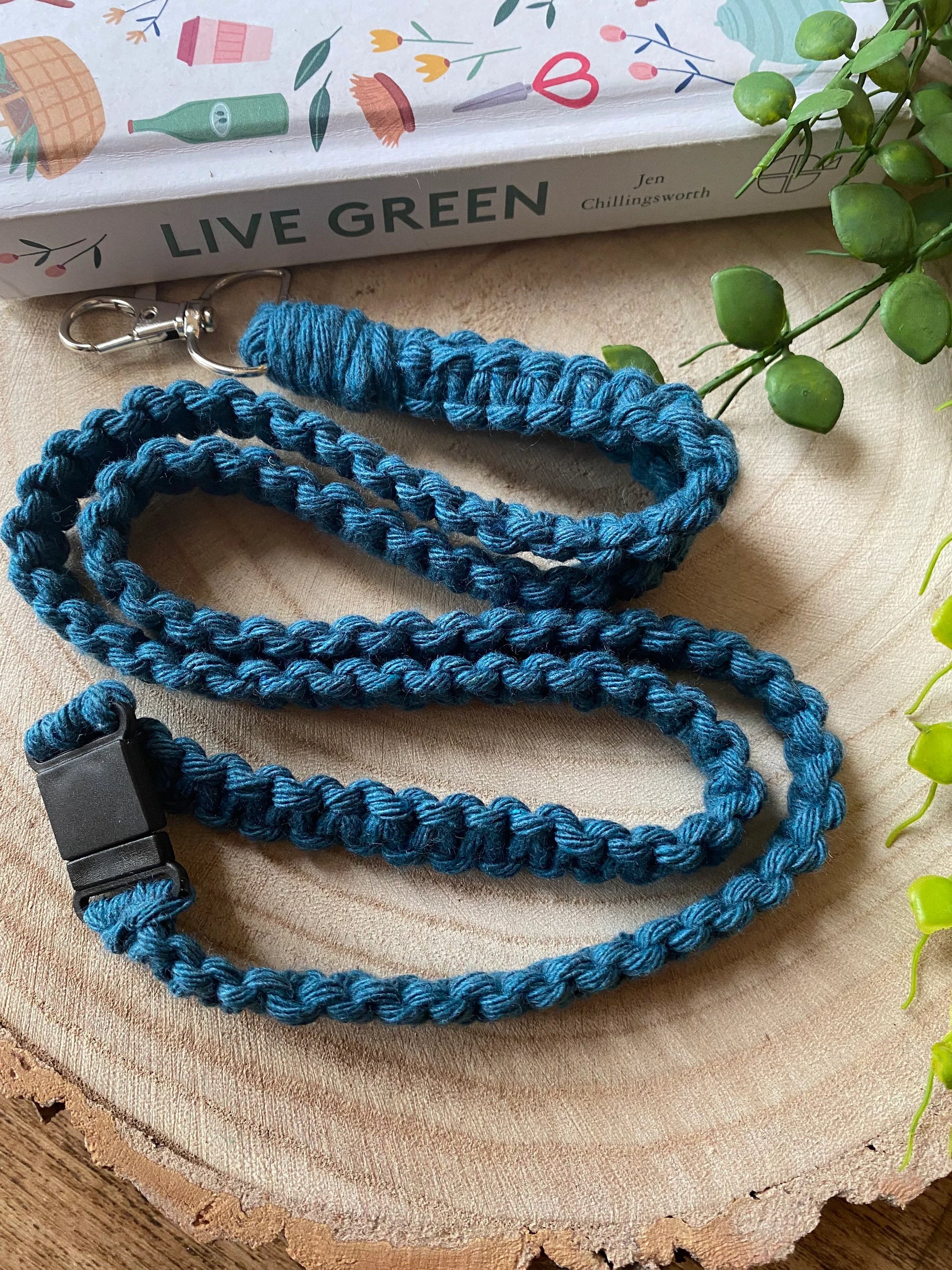 A handmade blue braided lanyard sits coiled on a wooden surface. There is a black safety break clip on one end and a metal clasp on the other. 