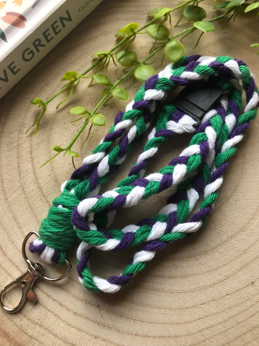 A handmade macrame lanyard badge holder with green, white, and purple strands lies coiled on a wooden surface. 