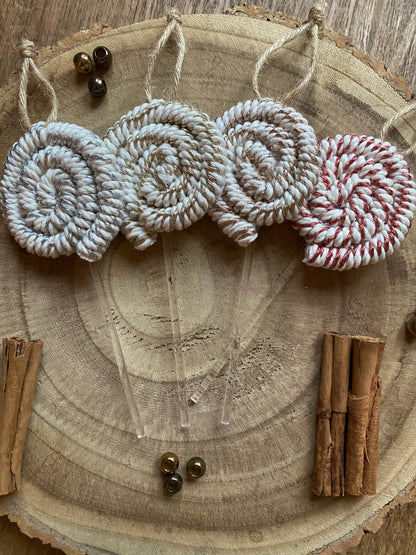 Four handmade macrame christmas decorations in the shape of lollipops, made with metallic yarn and clear acrylic sticks with a jute hanging loop at the top, displayed next to each other on a wooden board, surround by cinnamon sticks and gold beads for decoration. 