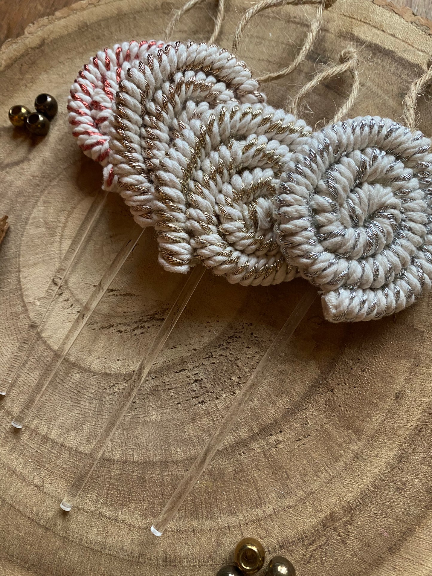 Four handmade macrame christmas decorations in the shape of lollipops, made with metallic yarn and clear acrylic sticks with a jute hanging loop at the top, displayed laying close to each other on a wooden board. 