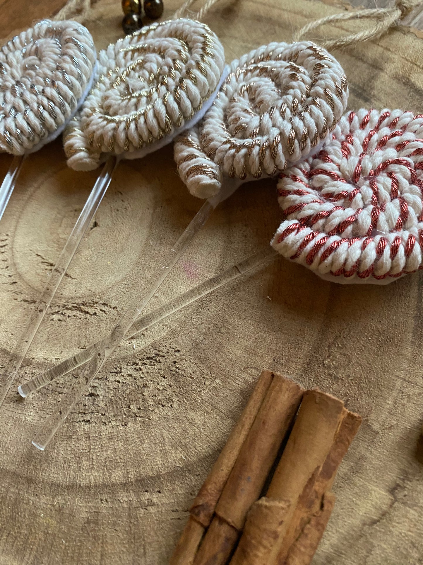 A set of four handmade macrame christmas decorations in the shape of lollipops, made with metallic yarn and clear acrylic sticks with a jute hanging loop at the top, displayed on a wooden surface next to a pile of cinnamon sticks. 
