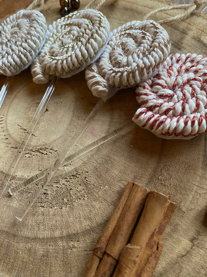 A set of four handmade macrame christmas decorations in the shape of lollipops, made with metallic yarn and clear acrylic sticks with a jute hanging loop at the top, displayed on a wooden surface next to a pile of cinnamon sticks. 