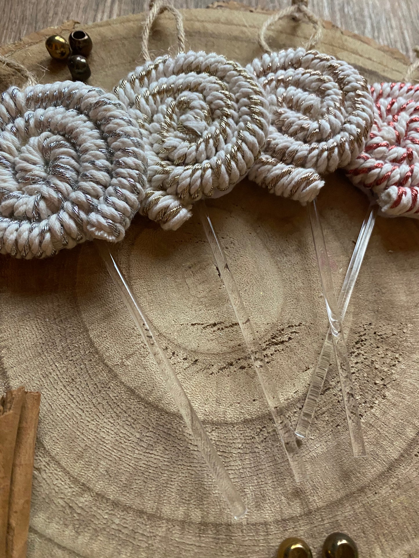 Close up of a set of four handmade macrame christmas decorations in the shape of lollipops, made with metallic yarn and clear acrylic sticks with a jute hanging loop at the top, displayed on a wooden surface next to a pile of cinnamon sticks. 