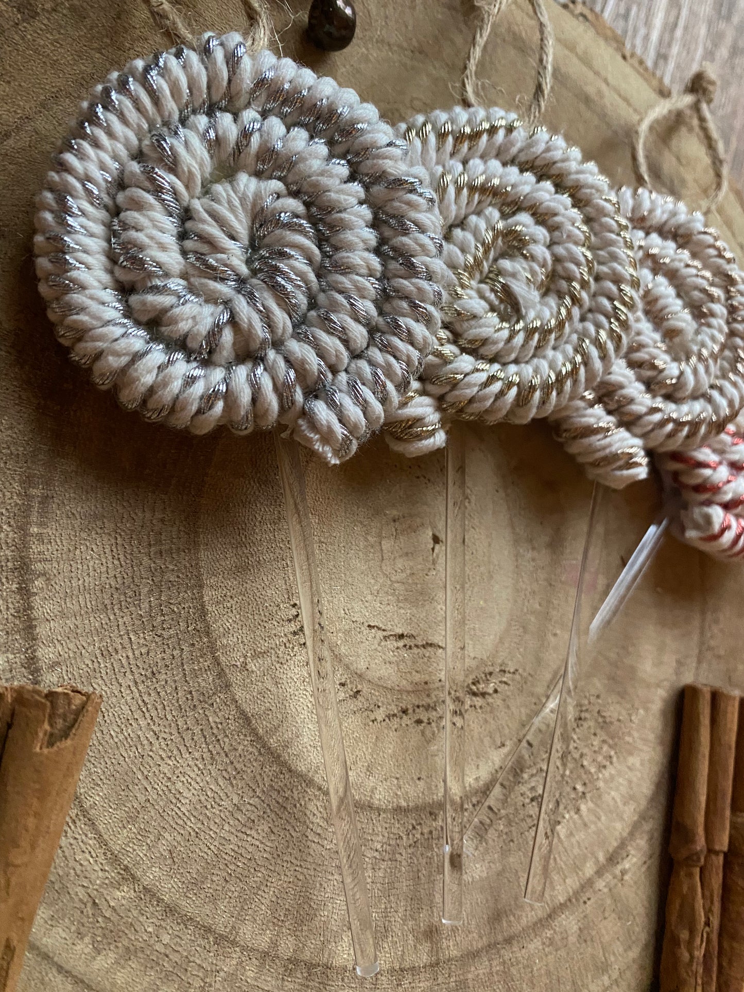 Close up image of a set of four handmade macrame christmas decorations in the shape of lollipops, made with metallic yarn and clear acrylic sticks with a jute hanging loop at the top, displayed on a wooden surface next to some cinnamon sticks. 