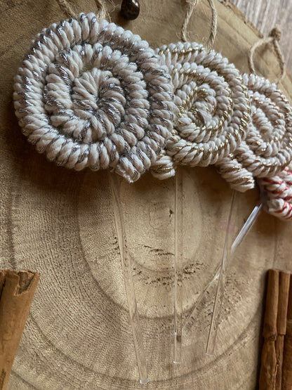 Close up image of a set of four handmade macrame christmas decorations in the shape of lollipops, made with metallic yarn and clear acrylic sticks with a jute hanging loop at the top, displayed on a wooden surface next to some cinnamon sticks. 