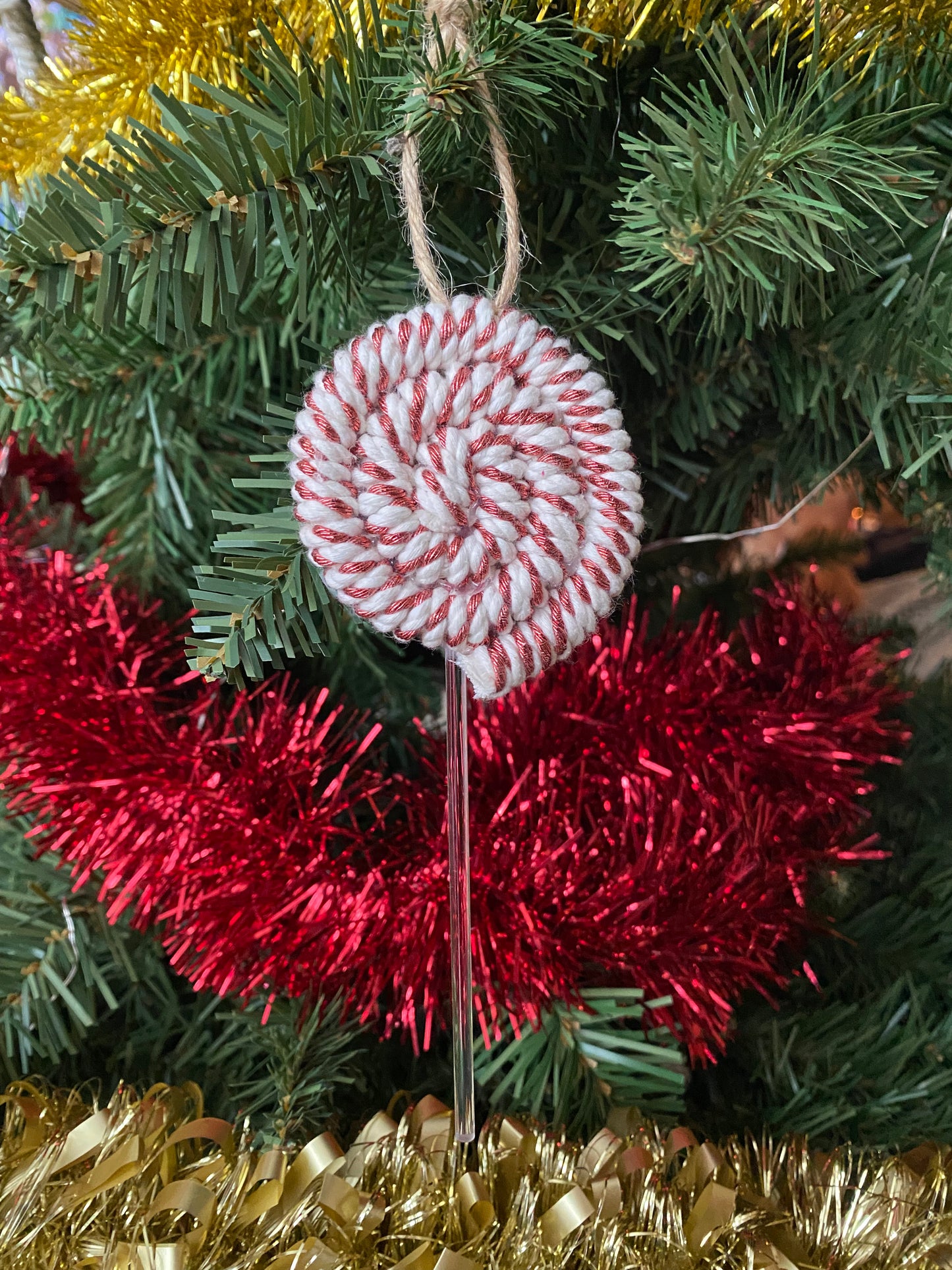 A handmade macrame christmas decoration in the shape of a lollipop, made with copper metallic yarn and clear acrylic stick with a jute hanging loop at the top, displayed hanging amongst red and gold tinsel and greenery on an artificial christmas tree. 