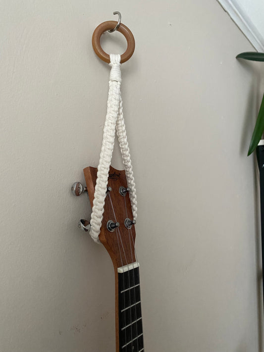 Close up of the headstock of a dark brown ukulele, hanging against a beige wall from a simple macrame knotted loop hanger, made with cotton yarn and a wooden ring. 