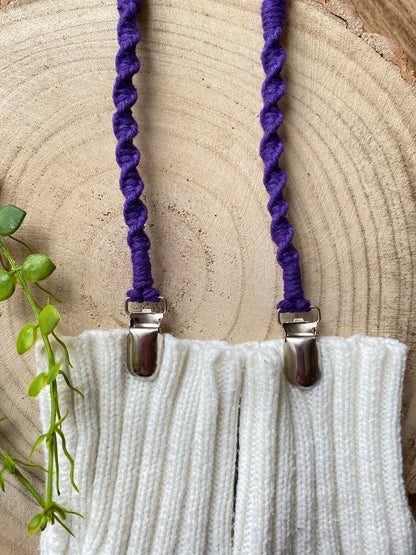 A pair of handmade macrame mitten clips made with purple yarn with metal clips on each end, attached to a pair of white mittens on a wooden board.